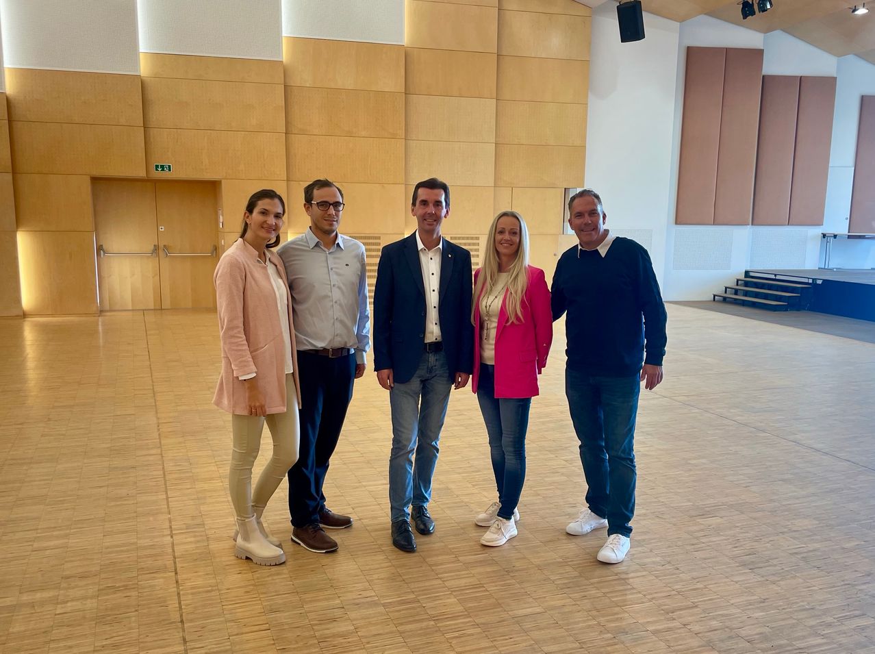 Fünf Menschen stehen in einer Reihe in einem großen Raum mit Holzböden und Wänden. Sie alle lächeln und posieren für ein Foto.