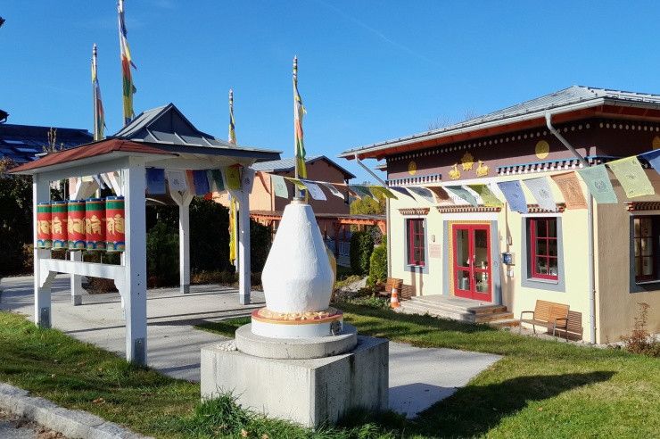 Ein tibetischer buddhistischer Tempel mit einem weißen Monument, Flaggen und einer roten Tür. In der Nähe befindet sich eine Bank und ein kleines Pavillon.