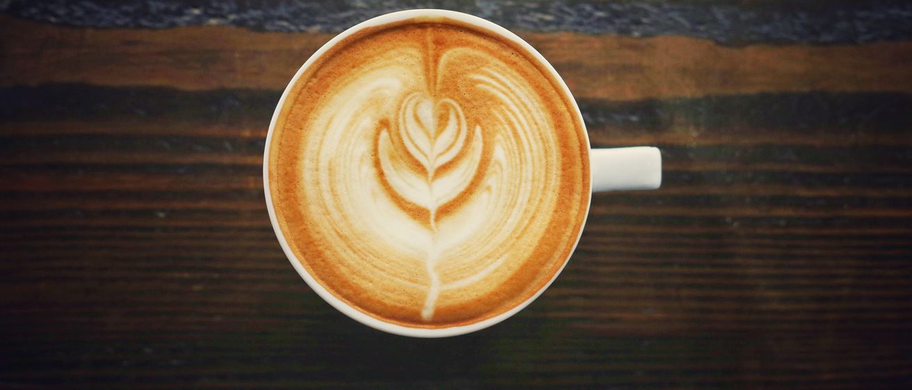 Eine Tasse Kaffee mit komplizierter Latte-Kunst, einem Herzdesign, steht auf einem hölzernen Tisch mit gestreiftem Hintergrund.