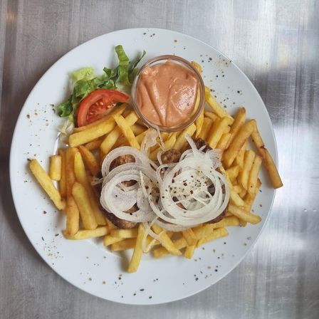 Ein weißer Teller mit Pommes, einem Fleischpäckchen mit Zwiebeln und einer Tasse Sauce auf einer Metalloberfläche.
