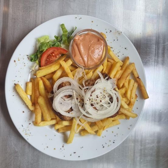 Ein weißer Teller mit Pommes, einem Fleischpäckchen mit Zwiebeln und einer Tasse Sauce auf einer Metalloberfläche.