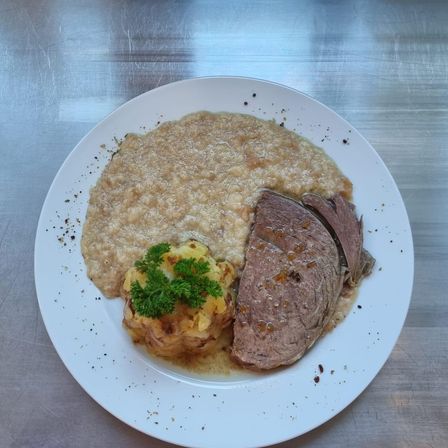 Ein weißer Teller mit einem Hauptgericht, bestehend aus geschnittenem Roastbeef mit Soße, Kartoffelpüree und Petersiliengarnitur. Der Teller liegt auf einer Metalloberfläche.