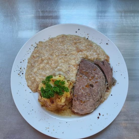 Ein weißer Teller mit einem Hauptgericht, bestehend aus geschnittenem Roastbeef mit Soße, Kartoffelpüree und Petersiliengarnitur. Der Teller liegt auf einer Metalloberfläche.