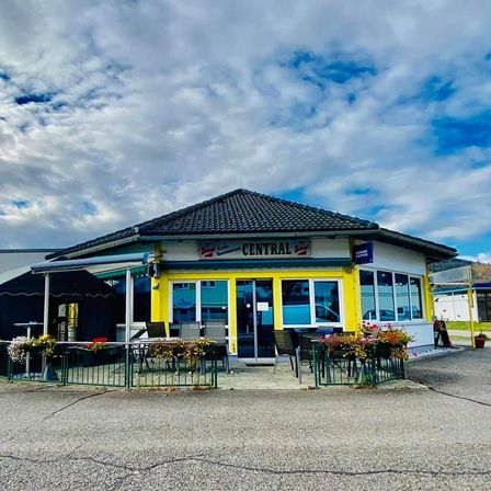 Ein helles gelbes und weißes Café namens Central, mit schwarzem Dach, Sitzgelegenheiten im Freien und einem klaren blauen Himmel darüber.