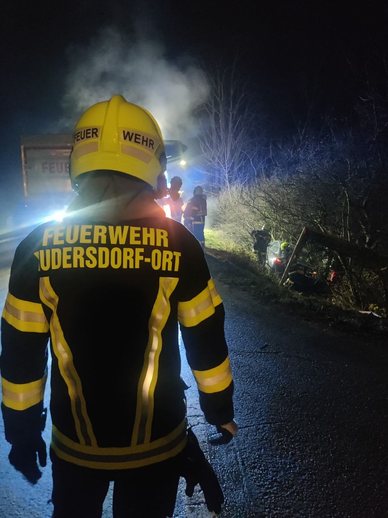 Ein Feuerwehrmann aus Udersdorf-Ort reagiert auf einen Unfall bei Nacht auf einer Straße, mit Rauch und Notfallleuchten sichtbar.
