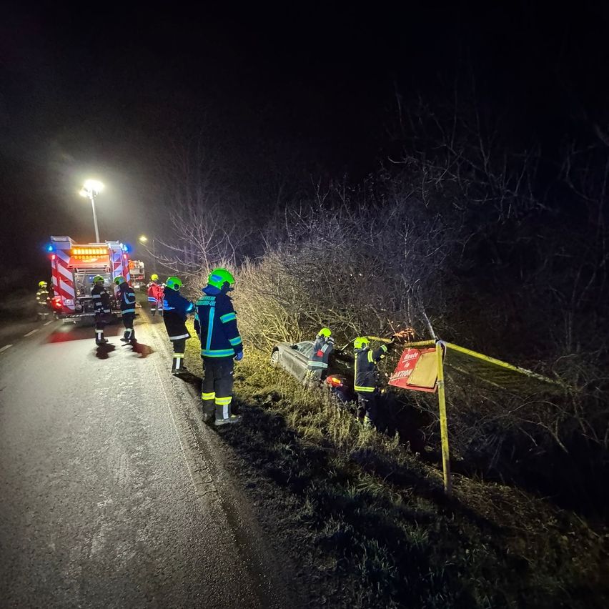 Einsatzkräfte in Schutzkleidung arbeiten nachts am Straßenrand. Ein Auto ist teilweise von der Straße abgekommen, und ein Feuerwehrwagen ist in der Nähe.