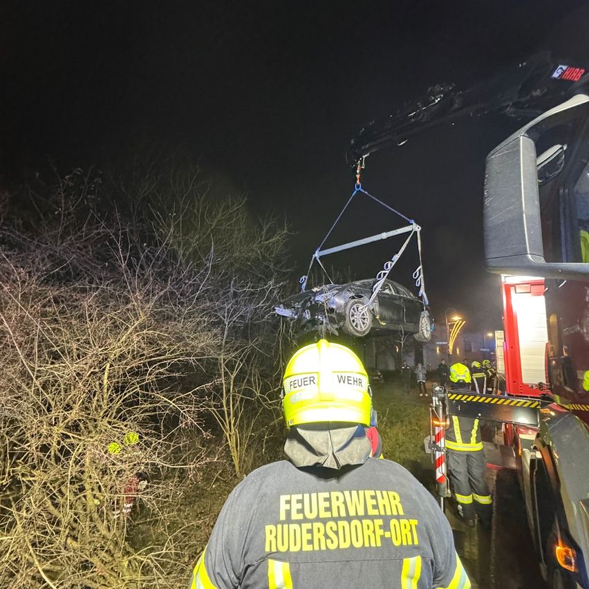 Feuerwehrleute heben ein demoliertes Auto mit einem Kran bei Nacht an. Das Auto ist an Seilen aufgehängt. Ein Feuerwehrmann in einem gelben Helm beaufsichtigt die Operation.