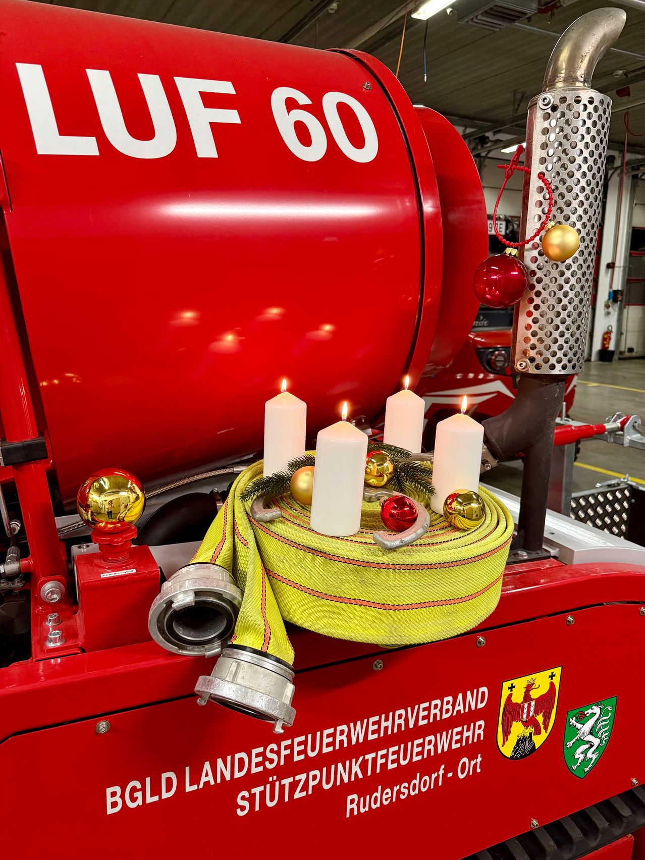 Ein roter Feuerwehrwagen mit der Aufschrift LUP 60, vier brennenden Kerzen, einem gelben Feuerwehrschlauch und Weihnachtsschmuck.
