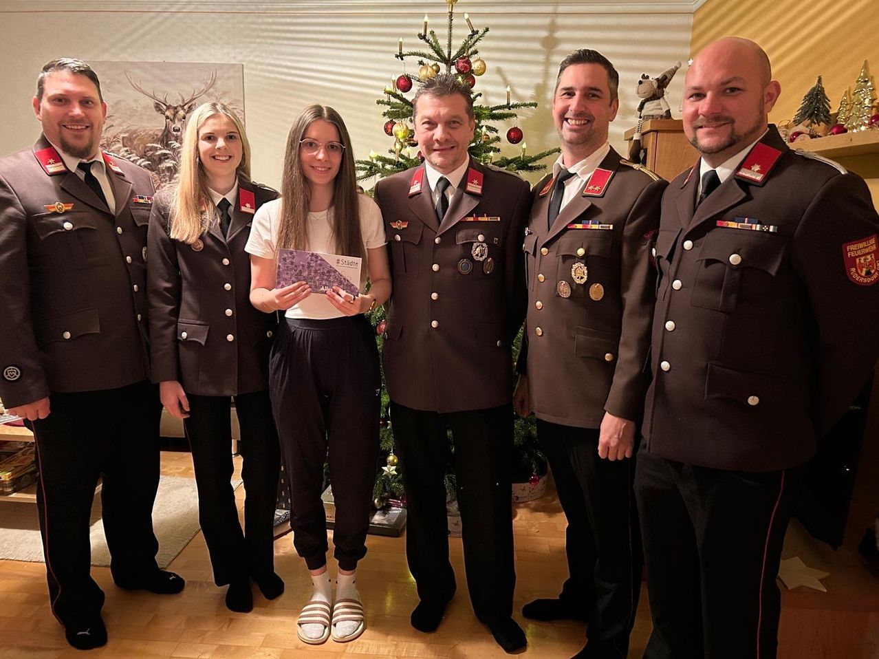 Fünf Personen, darunter vier in Militäruniformen, stehen vor einem Weihnachtsbaum. Eine Frau in der Mitte hält eine lila Karte. Dahinter ist ein Regal mit einem Plüschtier zu sehen.