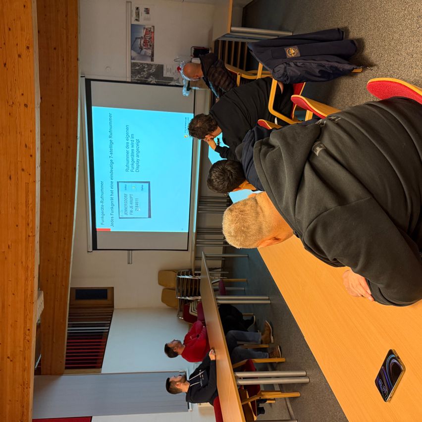 Mehrere Personen sitzen in einem Raum und beobachten eine Präsentation auf einer Projektionsleinwand. Eine Person steht vorne und zeigt auf den Bildschirm. Der Raum ist mit Stühlen, einem Tisch und einer Holzwand ausgestattet.