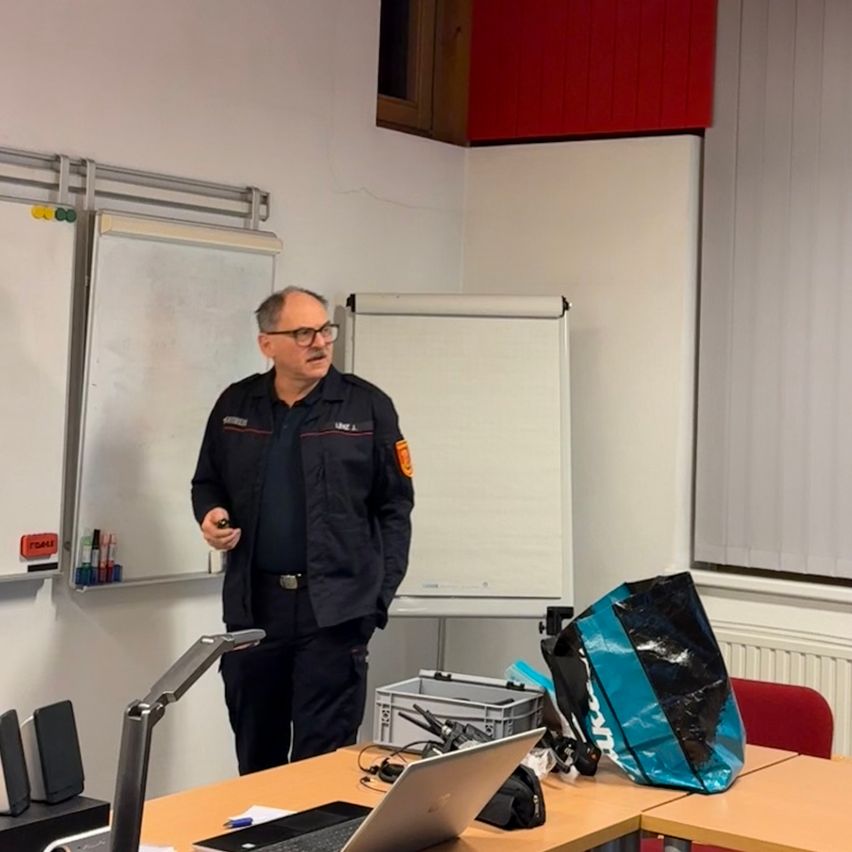 Ein Mann in Uniform steht in einem Klassenzimmer und hält eine Präsentation mit einer Fernbedienung. Eine Tafel und Marker sind an der Wand. Ein Laptop und ein blauer Beutel liegen auf dem Schreibtisch.