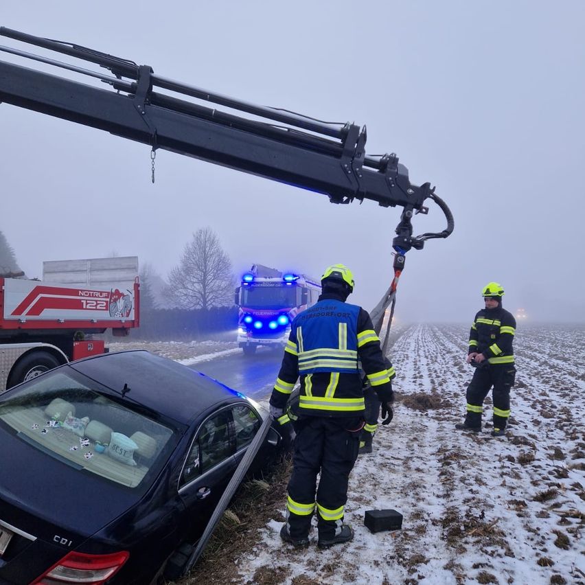 Feuerwehrleute benutzen einen Kran, um ein beschädigtes schwarzes Auto aus dem Schnee zu extrahieren. Ein Feuerwehrwagen steht hinter dem Auto, und ein anderer Feuerwehrmann steht in der Nähe.