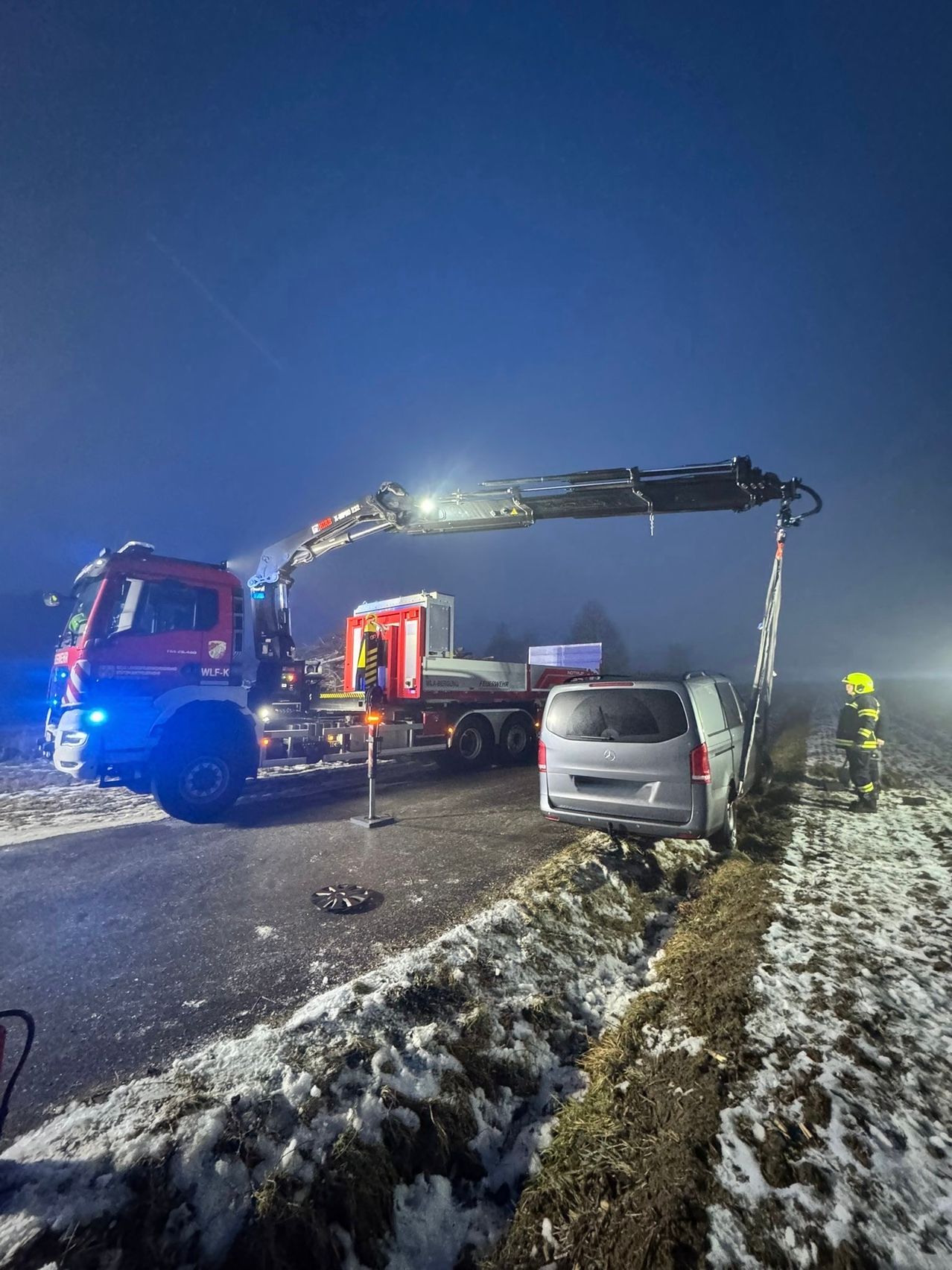 Ein Feuerwehrfahrzeug mit ausgeklapptem Kran steht neben einem grauen Van am Straßenrand, ein Feuerwehrmann steht in der Nähe. Die Szene spielt sich bei Nacht mit verschneiten Boden ab.