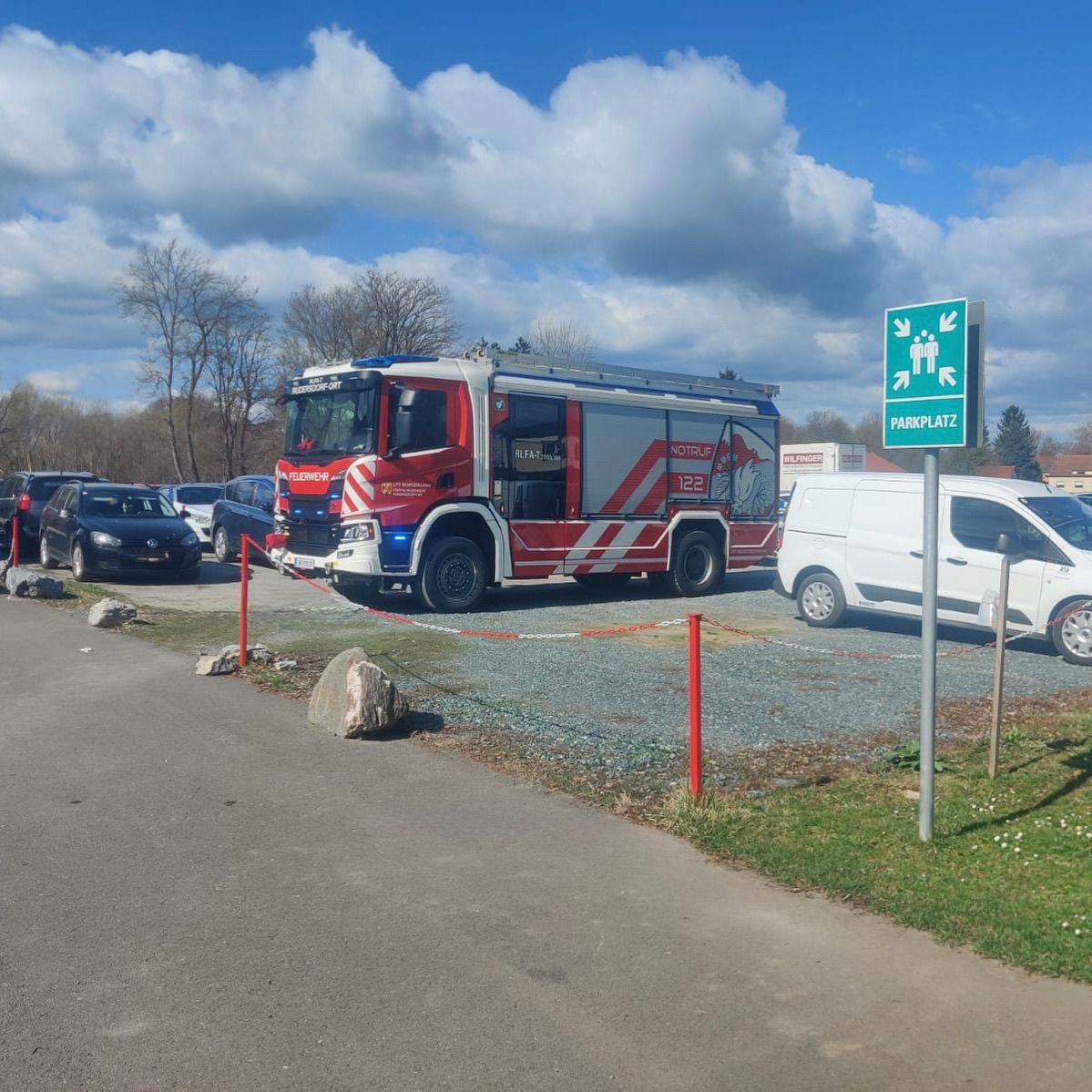 Ein Feuerwehrfahrzeug ist auf einem Parkplatz geparkt. Andere Autos sind darum herum geparkt. Ein weißer Van ist vor einem grünen Parkzeichen geparkt.