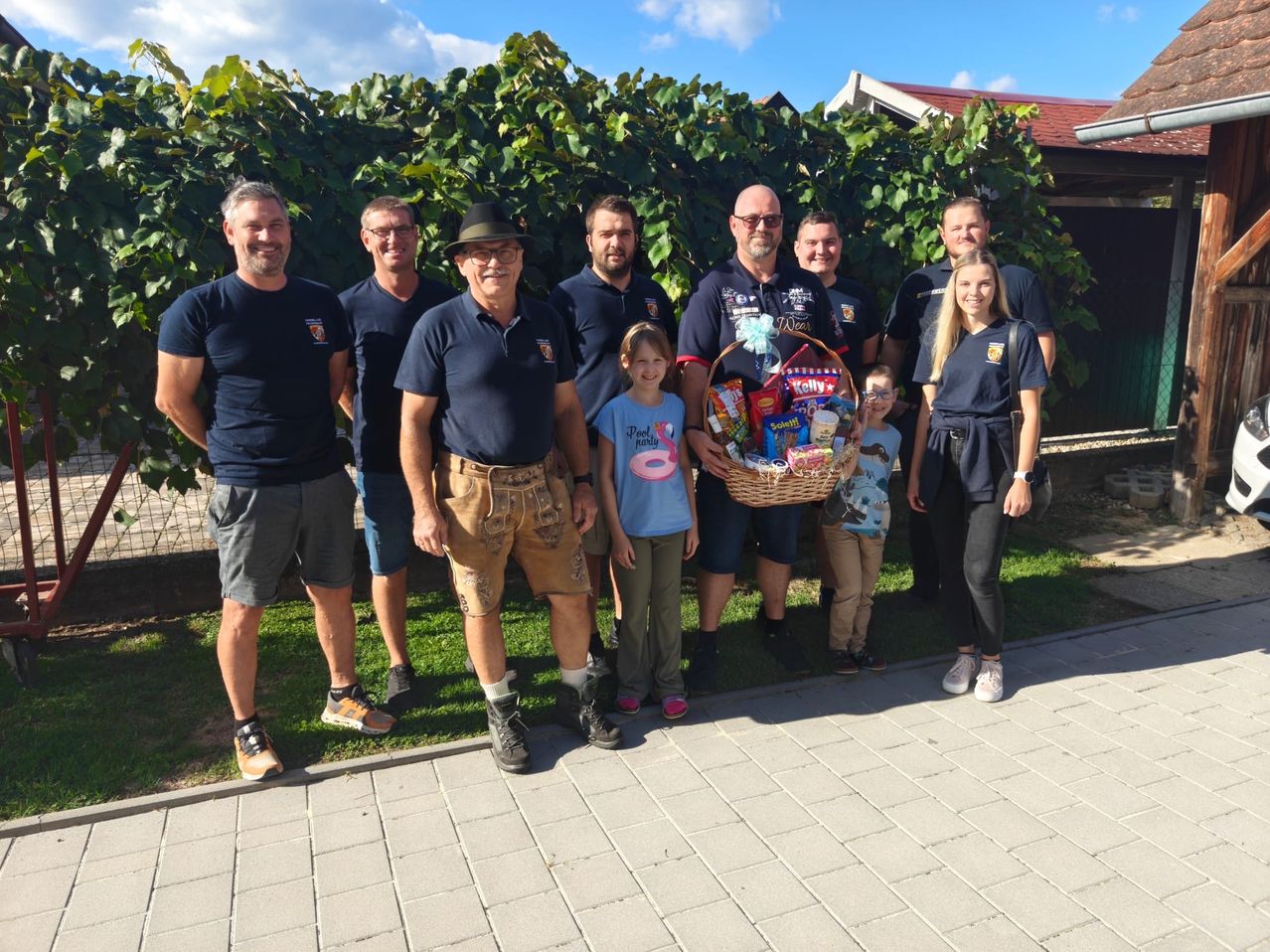A group of people, including adults and children, are standing together outside. They are smiling and posing for a photo. A man in the middle is holding a basket filled with snacks. Behind them, there are plants and a house.