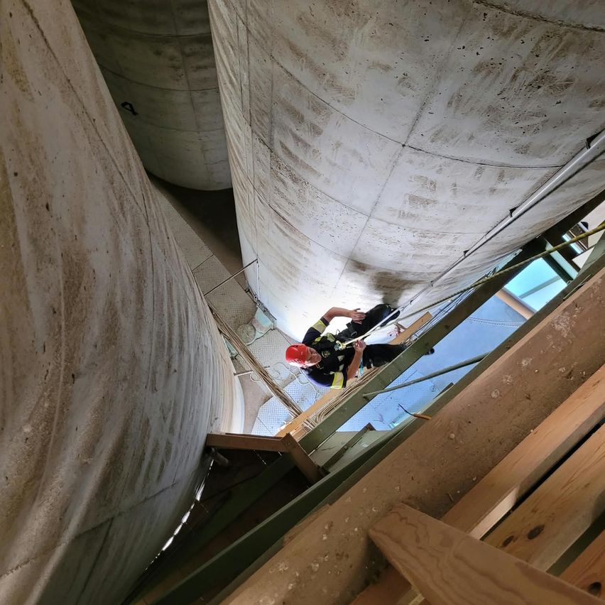 A firefighter in a red helmet is climbing up a large concrete structure. He is holding onto a rope and appears to be working on the ceiling.