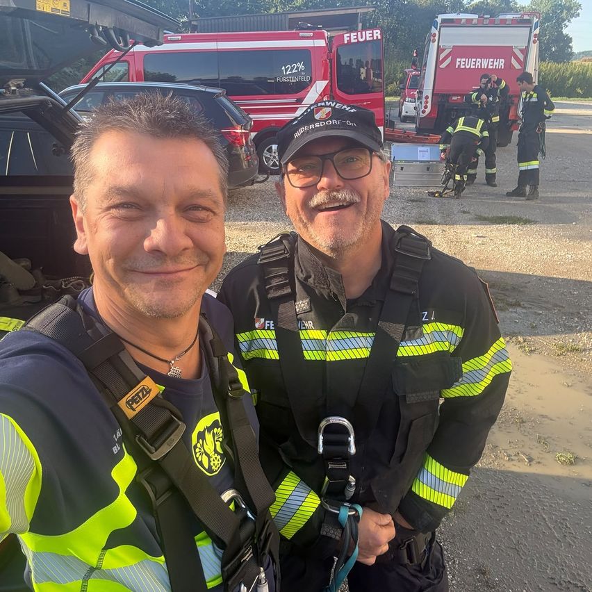 Two men in safety gear smile for a photo with a fire truck in the background.