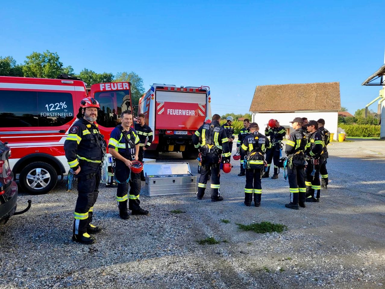 A group of firefighters in full gear are gathered around a fire truck. They are standing in a gravel area with trees and a house in the background.