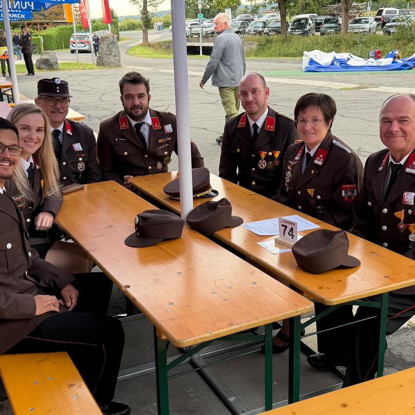 A group of uniformed individuals, possibly military personnel, are seated at a table with hats on it. They are smiling and posing for a photo. Behind them, there are parked cars and a person walking.