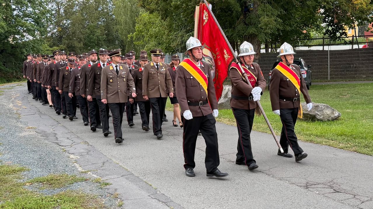 A group of military officers in uniform marching in a parade, carrying a flag and batons.
