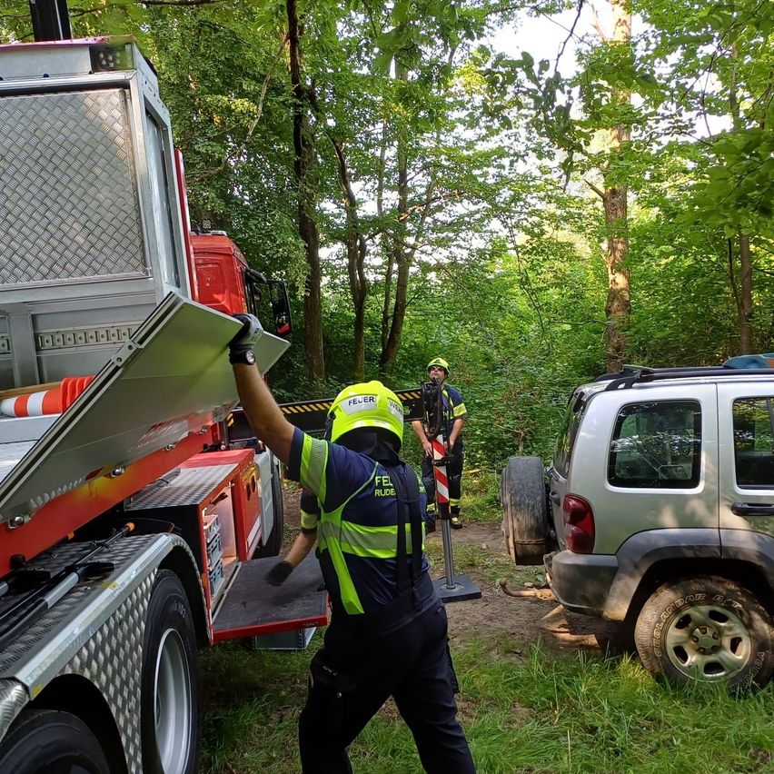 Zwei Feuerwehrleute entladen Ausrüstung von einem Lastwagen in einem bewaldeten Gebiet. Ein Feuerwehrmann trägt einen gelben Helm und eine reflektierende Weste, während ein anderer hinter einem geparkten SUV steht.