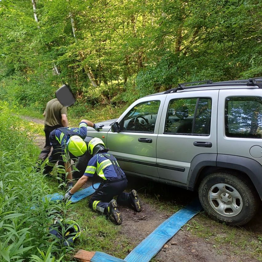 Eine Person mit Helm hilft einer anderen Person, beide in Sicherheitsausrüstung, um einen festsitzenden SUV in einem Wald. Das Fahrzeug hat ein Ersatzrad auf der Rückseite montiert.