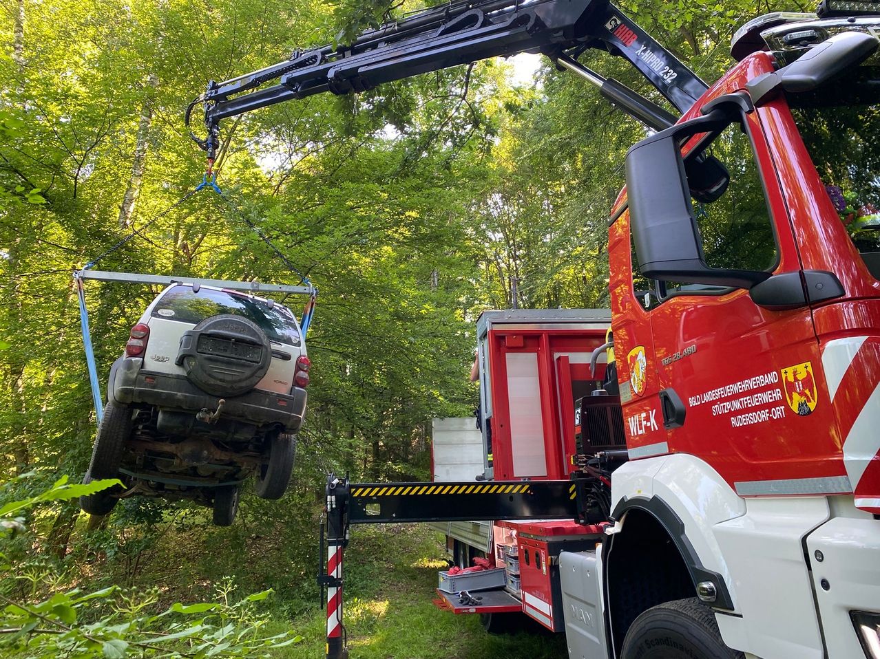 Ein Feuerwehrwagen mit angebrachtem Kran hebt ein Auto von der Erde, möglicherweise für eine Rettungsaktion. Das Fahrzeug wird durch eine Kette aufgehängt. Die Szene ist im Freien, mit Bäumen und Gras im Hintergrund.