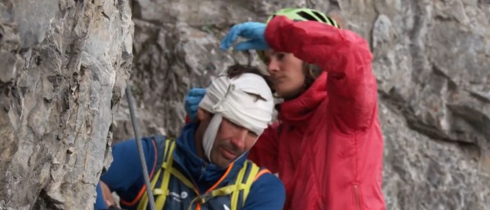 Drei Kletterer auf einem felsigen Berg. Einer hat einen weißen Verband am Kopf. Ein anderer trägt einen roten Anorak und einen Helm. Die dritte Person kniet mit einem blauen Helm.