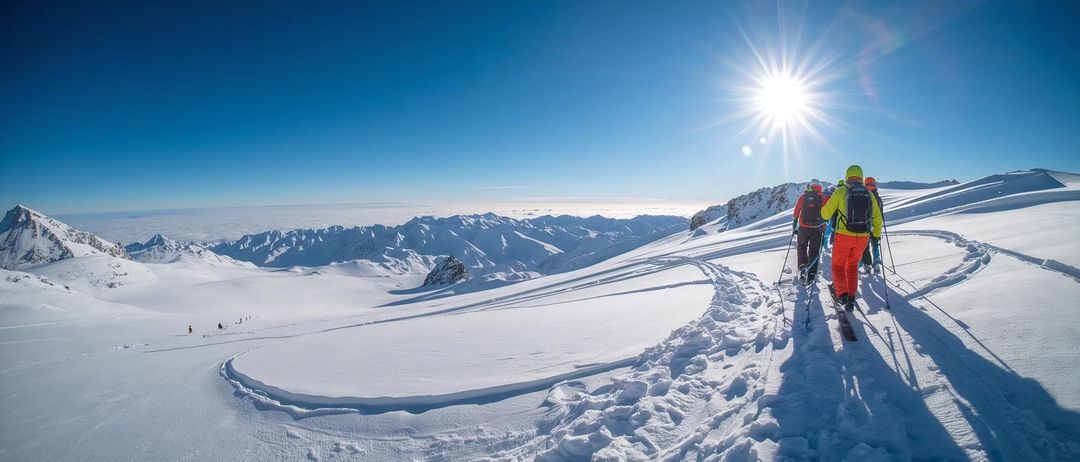 Zwei Personen skifahren einen schneebedeckten Berg hinauf. Die Sonne scheint hell am Himmel.