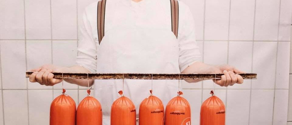 Ein Koch mit Hut und Brille hält einen Holzstab mit sechs orangefarbenen Würsten, die daran hängen. Die Würste sind mit weißen Schnüren gebunden.