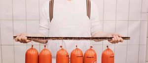 Ein Koch mit Hut und Brille hält einen Holzstab mit sechs orangefarbenen Würsten, die daran hängen. Die Würste sind mit weißen Schnüren gebunden.