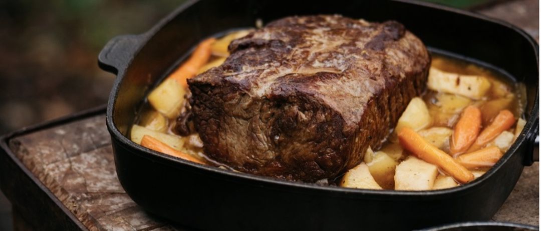 Ein schwarzer Topf mit gebratenem Fleisch und Gemüse steht auf einer Holzoberfläche. Daneben ist eine Schüssel mit Nudeln. Der Topf und die Schüssel stehen auf einem Holzkasten.