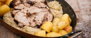 Auf einem Holztisch steht eine Kupferpfanne mit gebratenem Schweinefleisch, Sauerkraut und Kartoffeln. Das Fleisch ist verkohlt und in Scheiben geschnitten.