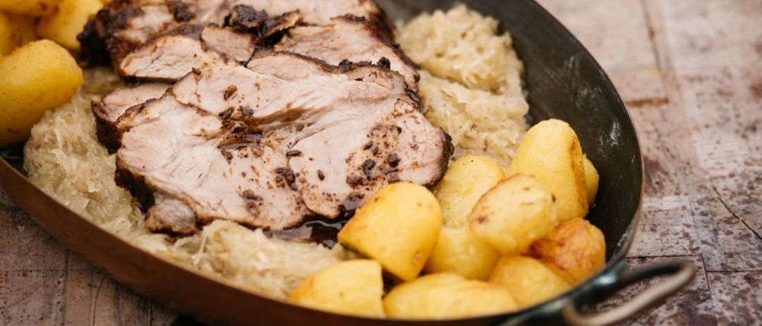 Auf einem Holztisch steht eine Kupferpfanne mit gebratenem Schweinefleisch, Sauerkraut und Kartoffeln. Das Fleisch ist verkohlt und in Scheiben geschnitten.