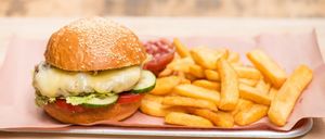 Ein Cheeseburger mit geschmolzenem Käse, Salat, Tomate und Gurke sitzt auf einem Brötchen mit Sesamkörnern. Er wird mit Ketchup und Pommes Frites serviert.