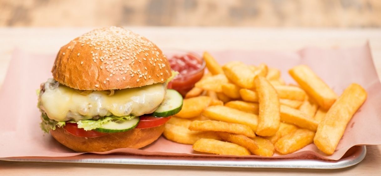 Ein Cheeseburger mit geschmolzenem Käse, Salat, Tomate und Gurke sitzt auf einem Brötchen mit Sesamkörnern. Er wird mit Ketchup und Pommes Frites serviert.