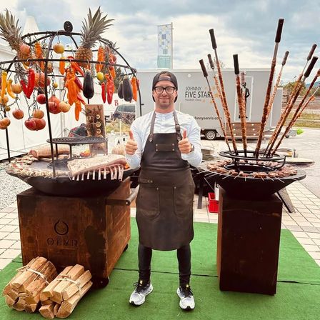 Ein Mann in einer Schürze steht vor einem mit Fleisch und Gemüse gefüllten Grill. Der Grill ist von Holzkisten und Holz umgeben, und er gibt einen Daumen nach oben.