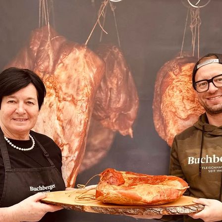 Zwei Menschen stehen vor einem Fleischerladenhintergrund und lächeln. Die Frau hält eine Holzplatte mit einem großen Stück Fleisch. Der Mann trägt eine Brille und eine Tarnjacke.