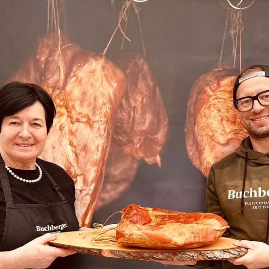 Zwei Menschen stehen vor einem Fleischerladenhintergrund und lächeln. Die Frau hält eine Holzplatte mit einem großen Stück Fleisch. Der Mann trägt eine Brille und eine Tarnjacke.