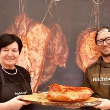 Zwei Menschen stehen vor einem Fleischerladenhintergrund und lächeln. Die Frau hält eine Holzplatte mit einem großen Stück Fleisch. Der Mann trägt eine Brille und eine Tarnjacke.