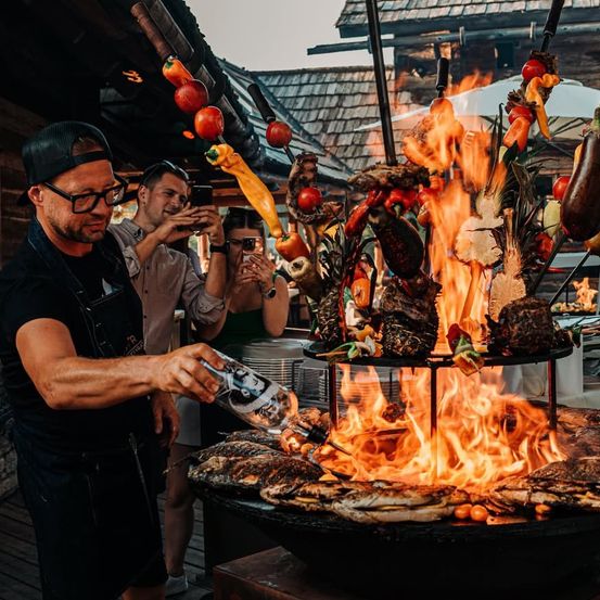 Ein Koch fügt einer brennenden Grillplatte bei einer Outdoor-Veranstaltung Sauce hinzu. Mehrere Personen machen Fotos. Verschiedene Fleisch- und Gemüsesorten werden auf Spießen gegrillt.