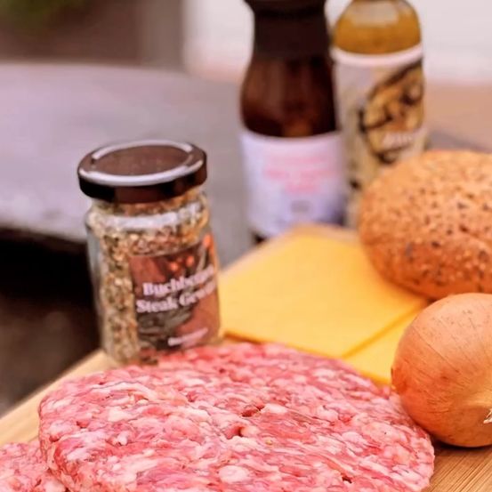 Ein Nahaufnahme von rohen Hamburger-Patties, begleitet von einem Glas Steakgewürz, Käsescheiben, einer Zwiebel und zwei Flaschen mit Saucen auf einem Holztisch.