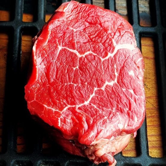 Ein roher Steak liegt auf einem schwarzen Grill mit einer hölzernen Oberfläche darunter und zeigt seine rote Farbe und feine Marmorierung.