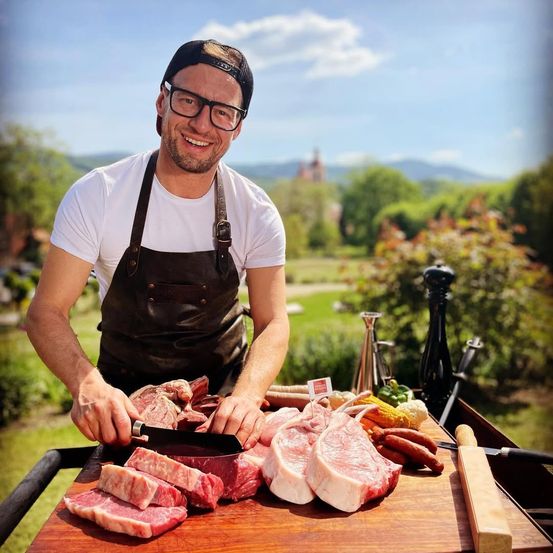 Ein Mann mit Brille und schwarzem Schürze bereitet Fleisch auf einem Holzschneidebrett in einer Außenumgebung vor.