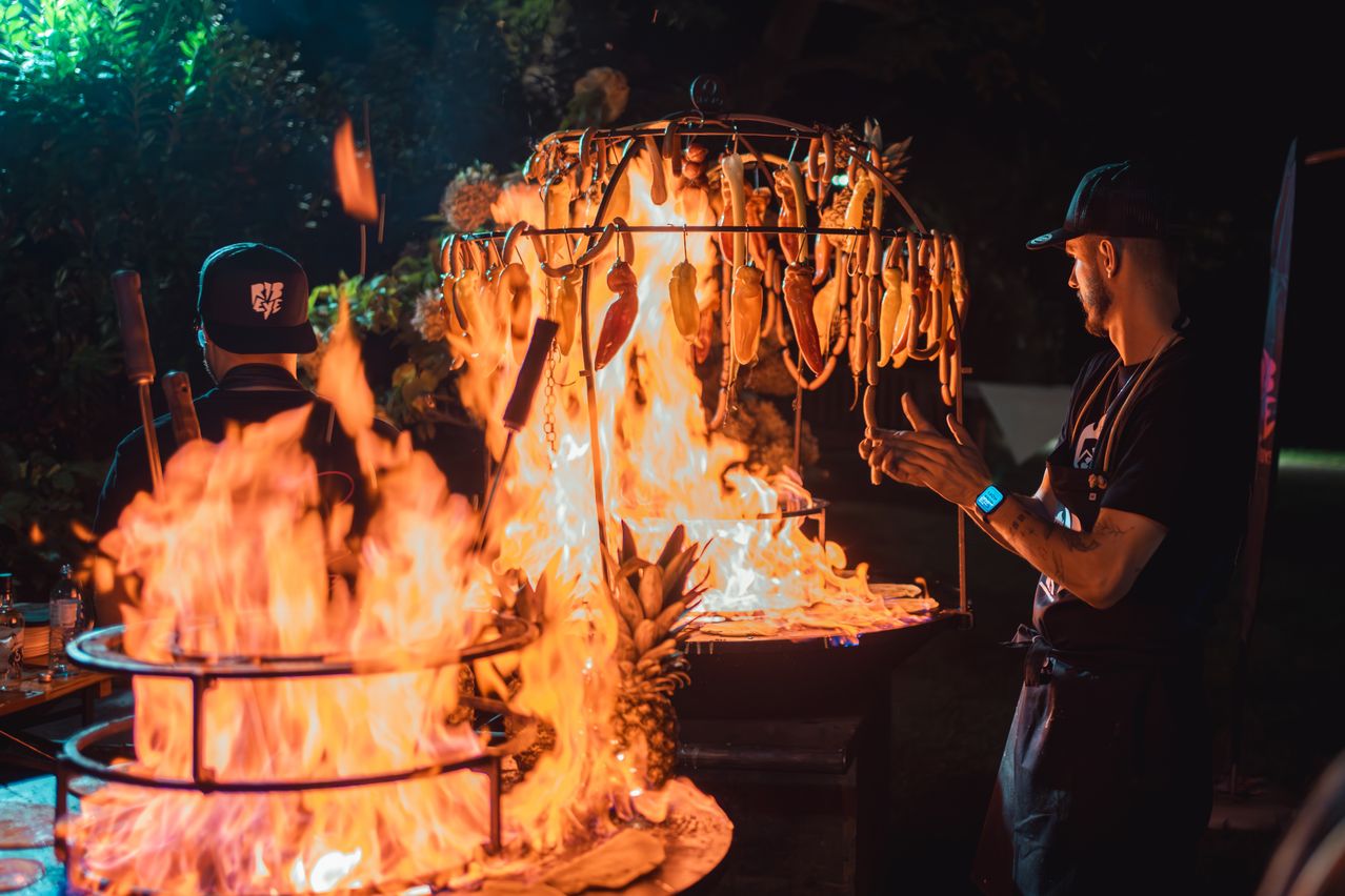 Ein Mann beaufsichtigt ein Grillfeuer mit großen Flammen und einer Ananas in der Nähe. Er trägt eine Kappe und eine Uhr. Eine andere Person in einer Kappe steht links.