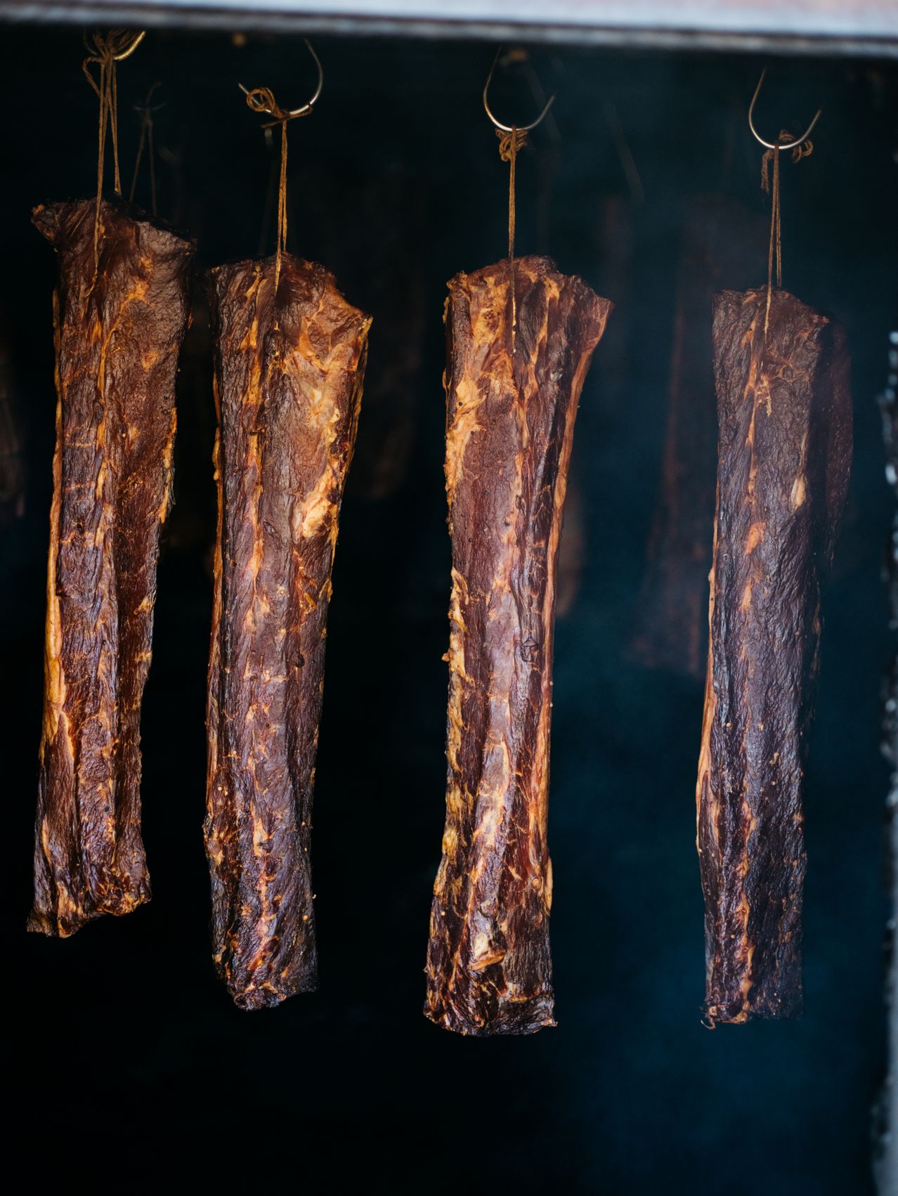 Vier Streifen geräuchertes Fleisch hängen an Schnüren in einem dunklen Raum. Das Fleisch hat eine knusprige Textur und eine rötlich-braune Farbe.