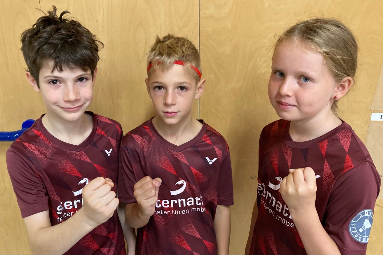 Drei junge Personen, zwei Jungen und ein Mädchen, posieren für ein Foto. Sie tragen burgunderrote Trikots mit weißen Logos. Der Junge in der Mitte trägt eine rote Stirnband. Sie alle machen Faustgesten. Hinter ihnen ist eine Holzwand.