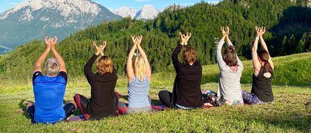 Fünf Personen sitzen in einer Reihe auf einem Grasfeld und praktizieren Yoga mit erhobenen Händen, unter einem klaren blauen Himmel mit Bergen in der Ferne.