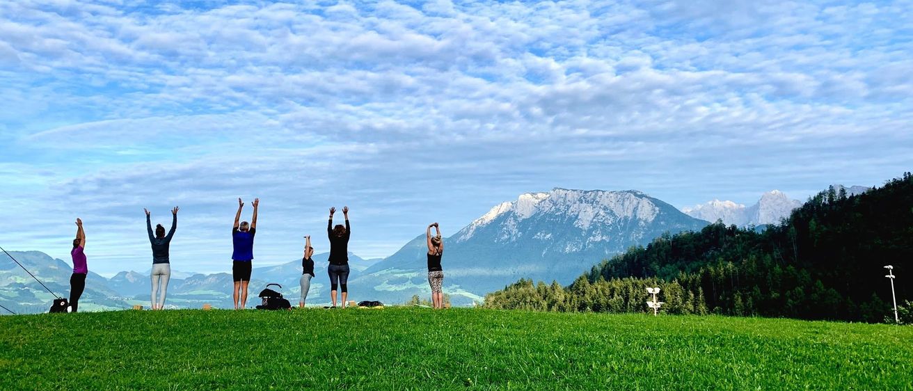 Eine Gruppe von Personen steht auf einer Wiese mit erhobenen Händen, möglicherweise macht Yoga, mit Bergen im Hintergrund.