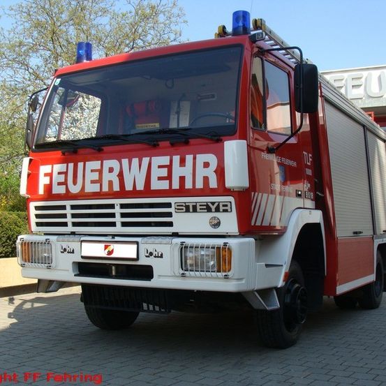 Ein rot-weißer Feuerwehrwagen mit der Aufschrift 'FEUERWEHR' und 'STEYR' steht vor einem Gebäude. Im Hintergrund befinden sich Büsche und Bäume.